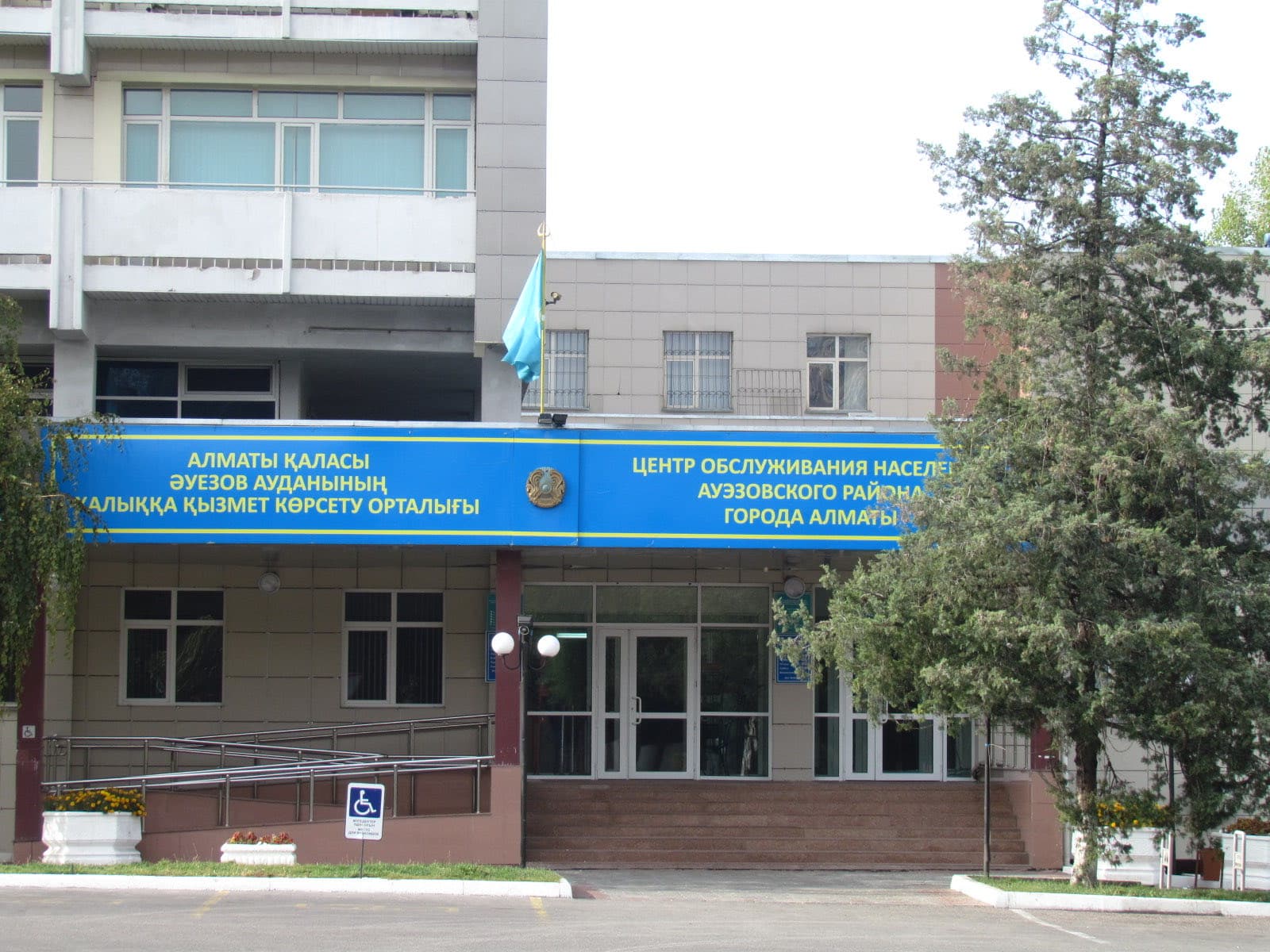 Вход в Центр обслуживания населения Ауэзовского района Алматы: синяя вывеска на казахском и русском над крыльцом, флаг Казахстана, пандус для колясок и ели у входа