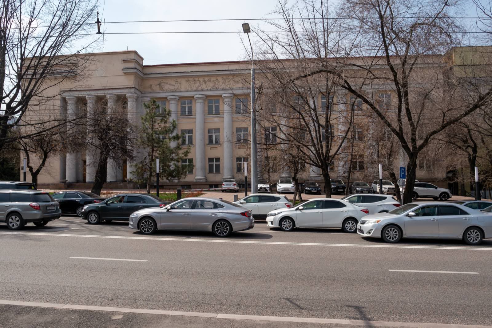 Проспект Абылай хана в Алматы: два ряда легковых машин у тротуара, за ними желтое здание с колоннами и голые весенние деревья