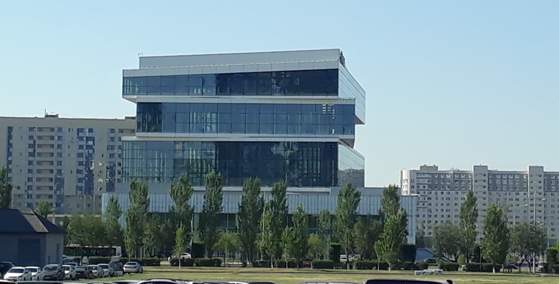 Стеклянный корпус ForteBank в Астане, перед ним газон с молодыми деревьями, по бокам панельные жилые дома